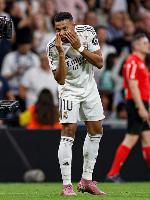 Jude Bellingham wearing a Real Madrid white jersey with number 10 and "Fly Emirates" text. He stands on a grass field, hands covering his face, wearing white socks and pink cleats. A referee in a red shirt and other players are visible in the background.