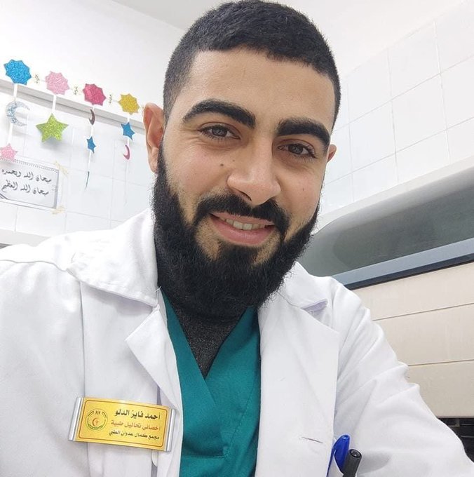Ahmad Faiz Al-Dalou wearing a white lab coat and green scrubs, standing in a tiled room with medical equipment. A name badge on his coat reads in Arabic. Decorative stars and Arabic text are visible on a bulletin board in the background.