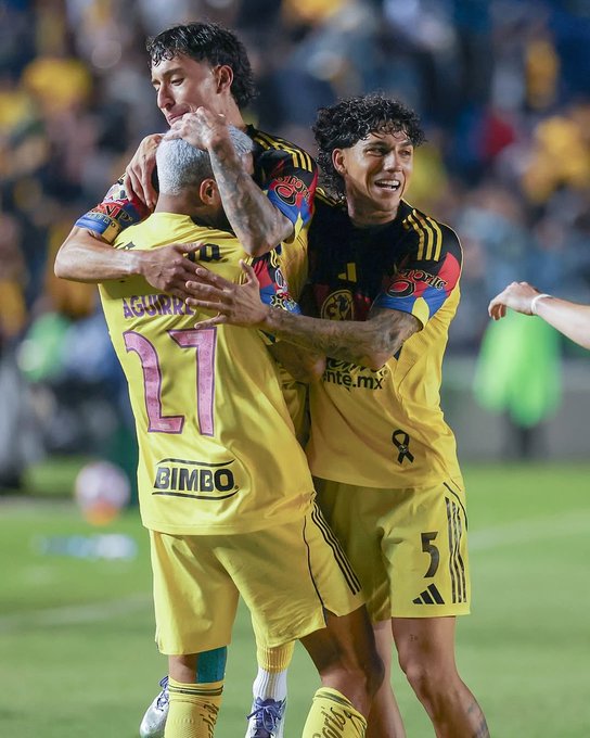 Tres jugadores de fútbol con camisetas amarillas con detalles en negro y rojo celebrando en un campo. Una camiseta tiene el nombre "Aguirre" y el número 27, otra tiene el número 5. Las camisetas muestran el logotipo "Bimbo" y el texto "América.mx". Los jugadores se abrazan, con una multitud visible en el fondo.