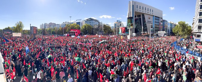 Large crowds holding Turkish and Palestinian flags, standing in an urban square with tall buildings. People carry banners with text in Arabic and Turkish, including "MUSLIMS UNITE" and "GAZZE İÇİN MEYDANLARDAYDIK." Some individuals wear traditional attire, and children hold signs reading "Gazze için dua ediyoruz."
