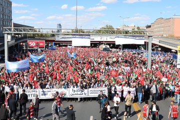 Large crowds holding Turkish and Palestinian flags, standing in an urban square with tall buildings. People carry banners with text in Arabic and Turkish, including "MUSLIMS UNITE" and "GAZZE İÇİN MEYDANLARDAYDIK." Some individuals wear traditional attire, and children hold signs reading "Gazze için dua ediyoruz."
