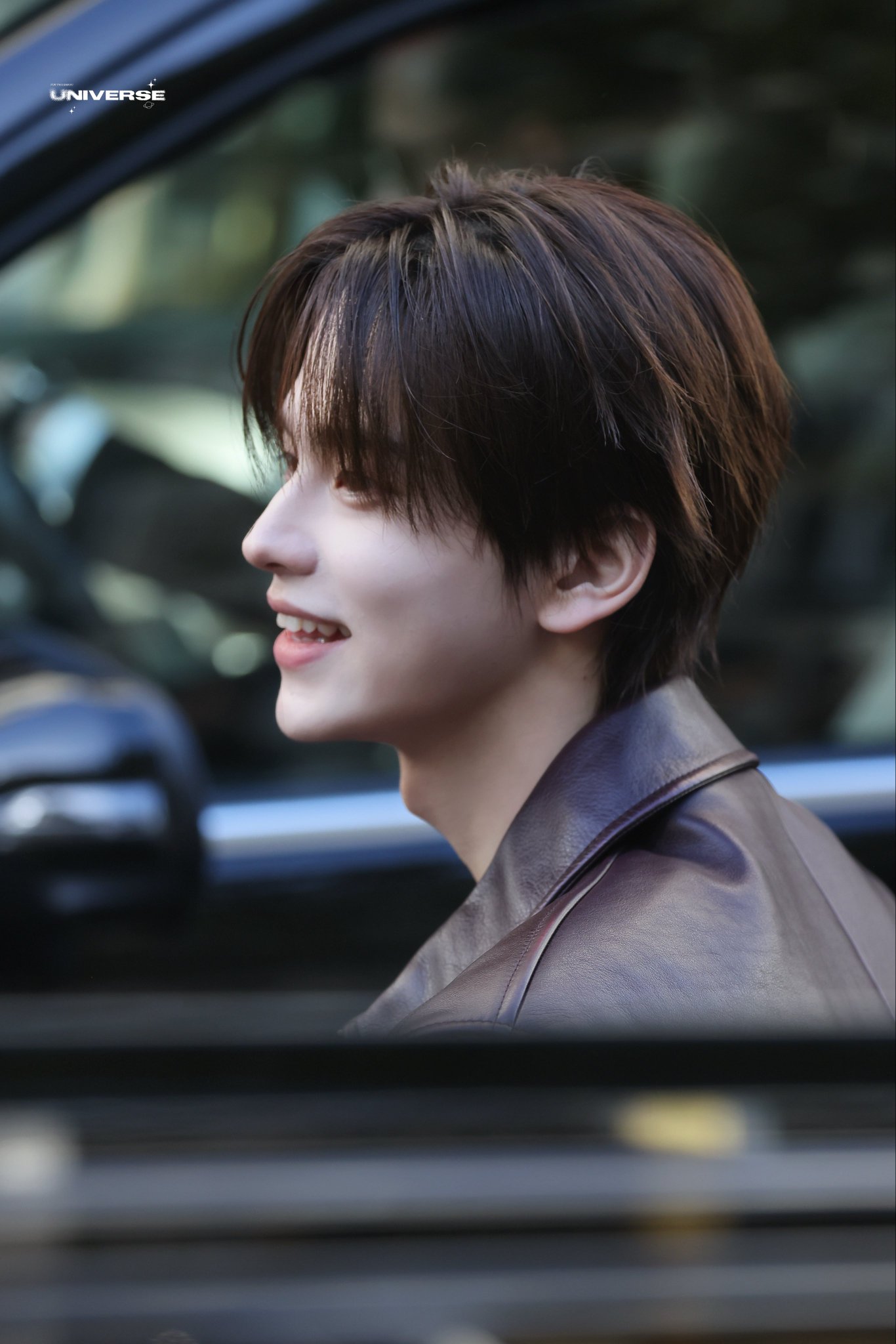Shin Yubin, a young man with dark brown hair, smiling softly in profile. He is wearing a dark leather jacket and is positioned near a car window, with greenery visible in the background. A watermark reading "UNIVERSE" is present in the upper left corner.