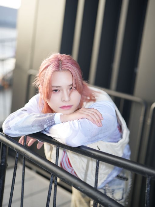 Matsuda Jin with pink hair, leaning on a black metal railing. He wears a light-colored shirt and vest, looking forward with a neutral expression. The background includes dark vertical panels and a blurred outdoor setting.