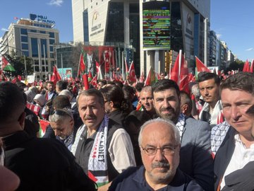 A large crowd of people gathered in an urban setting, holding numerous red and green flags with white symbols, including Turkish and Palestinian flags. Many individuals wear scarves and vests with text, some holding banners with Arabic and Turkish writing. The scene includes modern buildings, a mosque with minarets, and a clear blue sky.