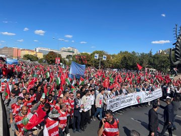 A large crowd of people gathered in an urban setting, holding numerous red and green flags with white symbols, including Turkish and Palestinian flags. Many individuals wear scarves and vests with text, some holding banners with Arabic and Turkish writing. The scene includes modern buildings, a mosque with minarets, and a clear blue sky.