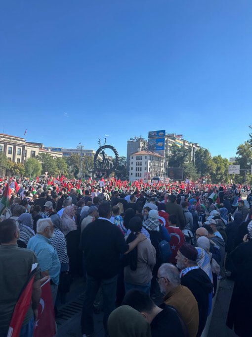 A large crowd of people gathered in an urban setting, holding numerous red and green flags with white symbols, including Turkish and Palestinian flags. Many individuals wear scarves and vests with text, some holding banners with Arabic and Turkish writing. The scene includes modern buildings, a mosque with minarets, and a clear blue sky.
