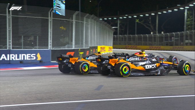 Lando Norris, Max Verstappen, and Oscar Piastri in Formula 1 cars on a racetrack during a night race. The cars, featuring orange and black McLaren designs with DHL and other sponsor logos, are closely positioned, showing contact on the first lap. The track is illuminated by overhead lights, with barriers and a grandstand visible in the background.