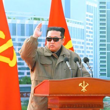 Kim Jong Un stands at a podium with two microphones, raising his right hand in a gesture. He wears sunglasses and a military-style jacket. Red flags with yellow symbols are behind him, and modern buildings are visible in the background. Benjamin Netanyahu sits at a desk with a microphone, wearing a suit and tie, with blue and white flags behind him.