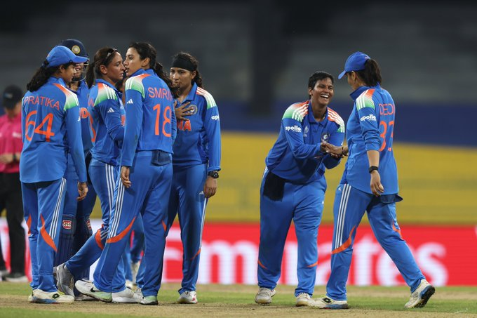 Indian women\'s cricket players in blue uniforms with orange accents, wearing jerseys with names like "HARMAN" and "REDDY" on the back, standing on a grassy field. They are shaking hands or celebrating together, with a stadium and yellow seats visible in the background. The players are wearing sports shoes and have their hair tied back.
