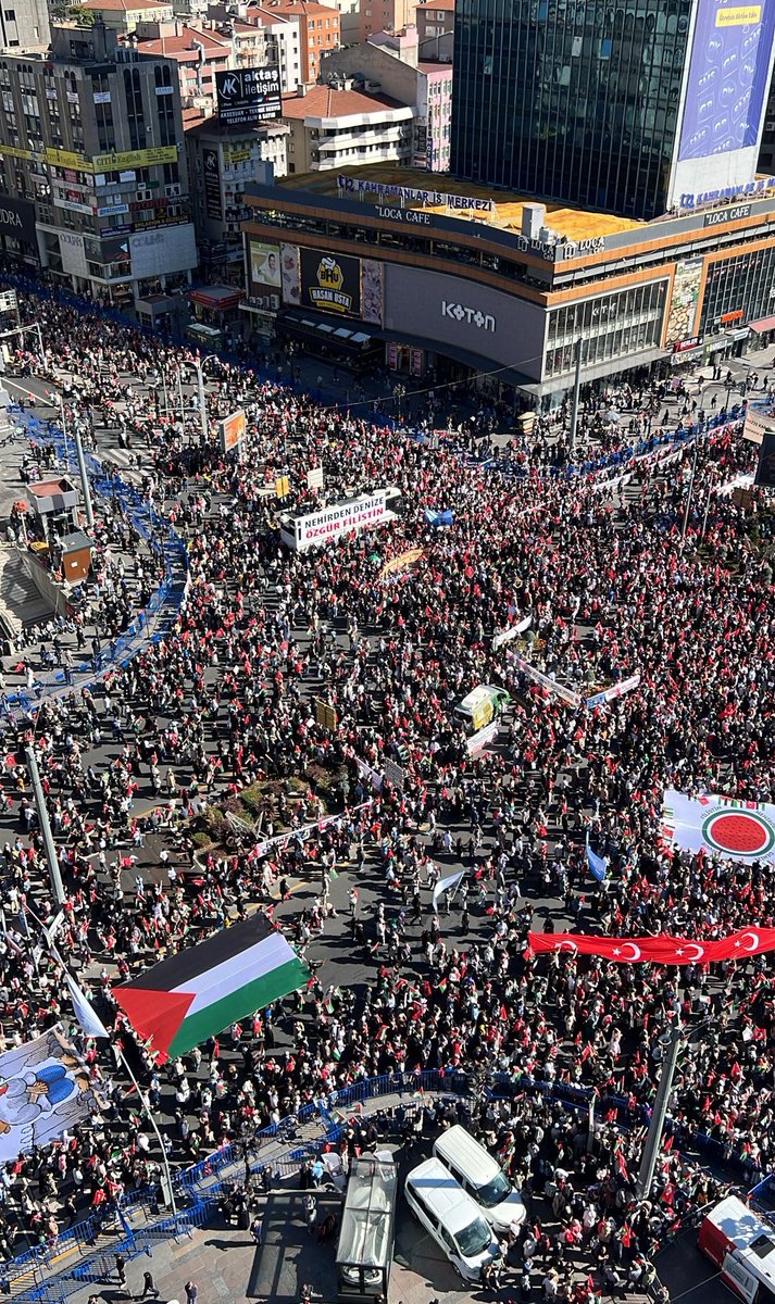 A large crowd of people gathered in an urban area, holding Palestinian flags and banners. The scene includes a prominent building with a digital billboard and vehicles in the background. Text on banners and flags is visible, including the Palestinian flag design.