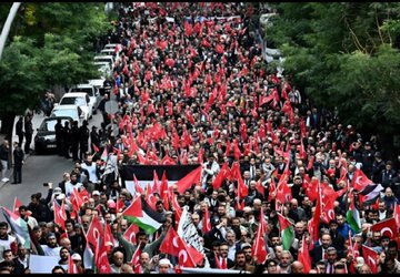 A large crowd gathered in an urban square holding Palestinian flags with red, green, white, and black colors. Many people wear keffiyehs and hold signs with text. The scene includes a prominent building and trees, with a clear sky overhead.