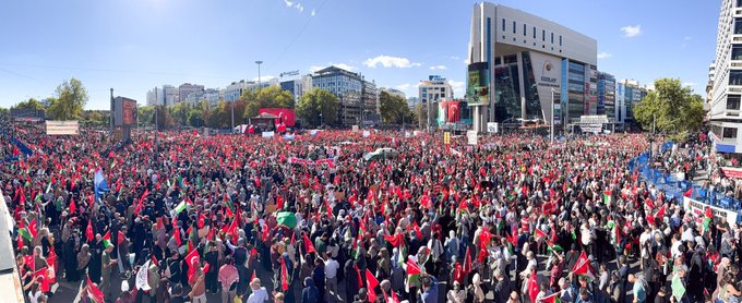 A large crowd gathered in an urban square holding Palestinian flags with red, green, white, and black colors. Many people wear keffiyehs and hold signs with text. The scene includes a prominent building and trees, with a clear sky overhead.