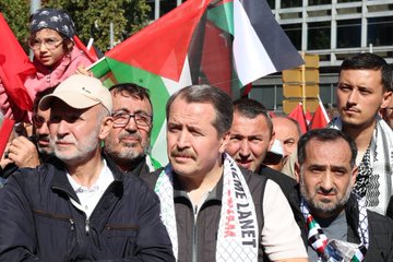 A large crowd gathered in an urban square holding Palestinian flags with red, green, white, and black colors. Many people wear keffiyehs and hold signs with text. The scene includes a prominent building and trees, with a clear sky overhead.