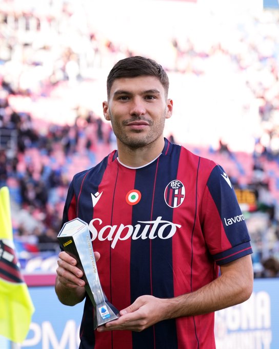 Nicolo Cambiaghi wearing a red and blue striped soccer jersey with the Bologna FC logo and Saputo sponsor text. He holds a trophy labeled Panini Player of the Match. A crowd is visible in the background.
