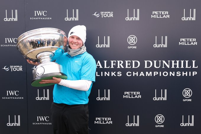 A person wearing a blue shirt, white long-sleeve undershirt, and white beanie holds a large silver trophy with a green base. The background features a black backdrop with logos for IWC Schaffhausen, Alfred Dunhill, Peter Millar, and the DP World Tour, along with text reading "Alfred Dunhill Links Championship."