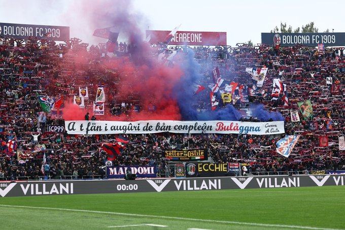 A crowded stadium filled with Bologna FC 1909 fans. Red and blue smoke rises from flares held by supporters. Banners display "Bologna FC 1909" and "A i fan i auguri di bu00e9n compleu00e0n Bulu00e0gna". The field is visible in the foreground with a scoreboard showing 00:00.