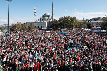 A large crowd holding Palestinian and Turkish flags gathers in Istanbul. People wave flags and hold a banner reading "FİLİSTİN ÖZGÜR OLANA DEK" in red and black text. The background features the Ayasofya mosque with its distinctive domes and minarets.