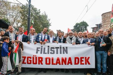 A large crowd holding Palestinian and Turkish flags gathers in Istanbul. People wave flags and hold a banner reading "FİLİSTİN ÖZGÜR OLANA DEK" in red and black text. The background features the Ayasofya mosque with its distinctive domes and minarets.