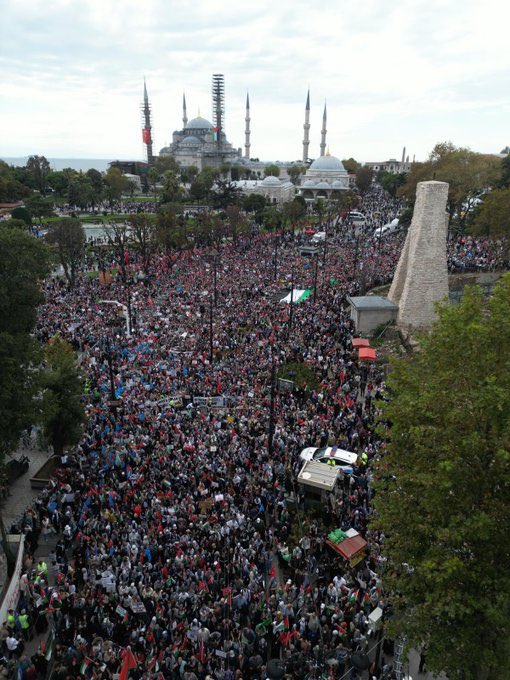 A large crowd holding Palestinian and Turkish flags gathers in Istanbul. People wave flags and hold a banner reading "FİLİSTİN ÖZGÜR OLANA DEK" in red and black text. The background features the Ayasofya mosque with its distinctive domes and minarets.
