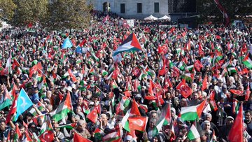 A large crowd holding Palestinian and Turkish flags gathers in Istanbul. People wave flags and hold a banner reading "FİLİSTİN ÖZGÜR OLANA DEK" in red and black text. The background features the Ayasofya mosque with its distinctive domes and minarets.