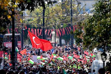 A large crowd of people gathered in an outdoor urban setting, holding numerous red and green flags with white and black stripes, resembling the Turkish and Palestinian flags. Many individuals raise their hands, some holding signs. Trees, a bus, and a historic building with domes and minarets, likely a mosque, are visible in the background. Text on a sign reads "Fatih."
