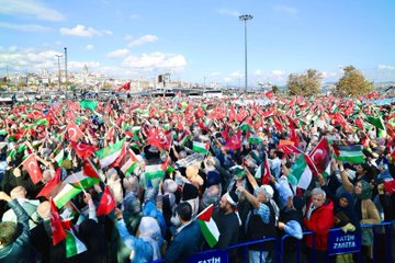 A large crowd of people gathered in an outdoor urban setting, holding numerous red and green flags with white and black stripes, resembling the Turkish and Palestinian flags. Many individuals raise their hands, some holding signs. Trees, a bus, and a historic building with domes and minarets, likely a mosque, are visible in the background. Text on a sign reads "Fatih."