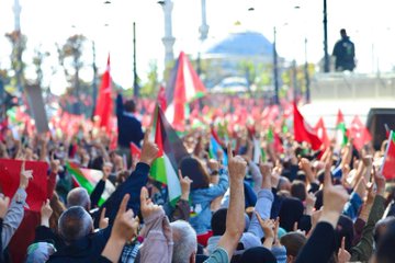A large crowd of people gathered in an outdoor urban setting, holding numerous red and green flags with white and black stripes, resembling the Turkish and Palestinian flags. Many individuals raise their hands, some holding signs. Trees, a bus, and a historic building with domes and minarets, likely a mosque, are visible in the background. Text on a sign reads "Fatih."