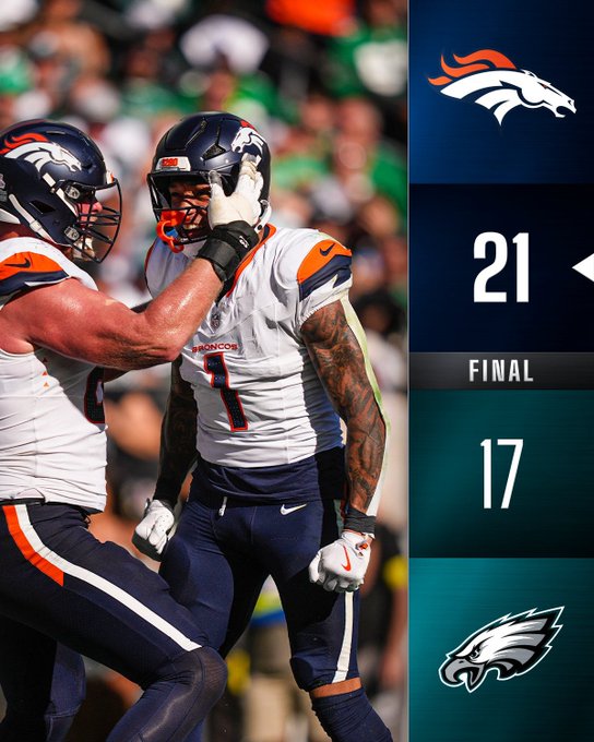 Dos jugadores de los Denver Broncos con uniformes blancos y azul marino con detalles en naranja, con cascos, celebrando en un campo de fútbol. Un jugador tiene el número 1 en su camiseta y tatuajes visibles en los brazos. El marcador muestra un marcador final de 21-17, con el logotipo de los Denver Broncos y el logotipo de los Philadelphia Eagles.