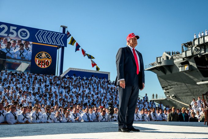 Donald Trump stands on a platform wearing a dark suit, red tie, and red cap. Behind him, a large crowd of people in white uniforms is visible, seated in bleachers. A massive aircraft carrier is docked nearby, and a large sign reading "250" with an emblem is displayed. Blue and yellow flags hang above the crowd.