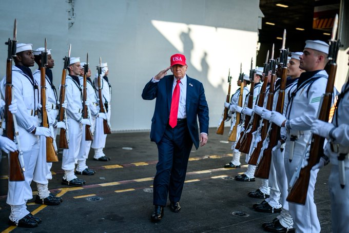 Donald Trump wearing a red USA cap and red tie, walking forward in a blue suit. He is flanked by rows of uniformed sailors in white holding rifles, standing in formation on a deck with a large white wall in the background.