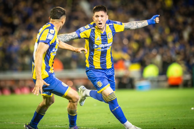 Two soccer players in blue and yellow striped jerseys celebrating on a field. One player has tattoos on his arms and is raising a fist, while the other extends an arm toward him. The background shows a stadium with a crowd and a grassy pitch.