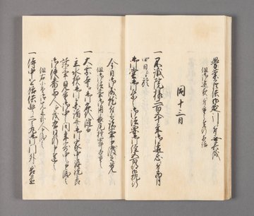 Open aged booklet with Japanese calligraphy on cream paper showing detailed black ink characters arranged in vertical columns across both pages describing historical event details in classical script style. Second image displays another open page of similar booklet with intricate vertical text in black ink on aged paper capturing formal records of locations and procedures.