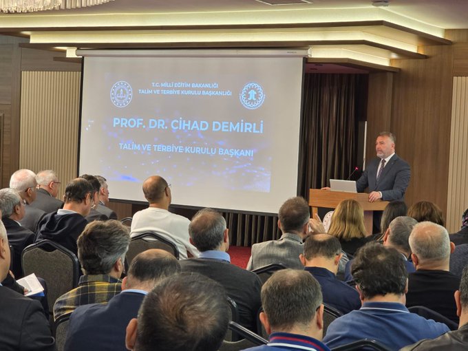 A conference room with attendees seated in rows, facing a speaker at a podium. A projection screen displays text reading "Talim ve Terbiye Kurulu Başkanlığı" and "Eğitim Politikaları Daire Başkanlığı Denklik, Belge Doğrulama İşlemleri ve e-Denklik Modülü Eğitimi Semineri" with logos. The speaker, a man in a suit, stands addressing the audience. A Turkish flag is visible on a banner in the background.