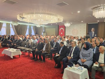 A conference room with attendees seated in rows, facing a speaker at a podium. A projection screen displays text reading "Talim ve Terbiye Kurulu Başkanlığı" and "Eğitim Politikaları Daire Başkanlığı Denklik, Belge Doğrulama İşlemleri ve e-Denklik Modülü Eğitimi Semineri" with logos. The speaker, a man in a suit, stands addressing the audience. A Turkish flag is visible on a banner in the background.