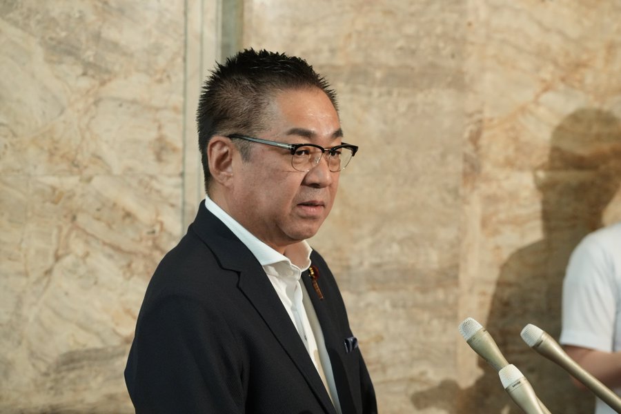 Endo Takashi wearing a dark suit, white shirt, and glasses, standing in front of microphones. A textured beige background is visible behind him.