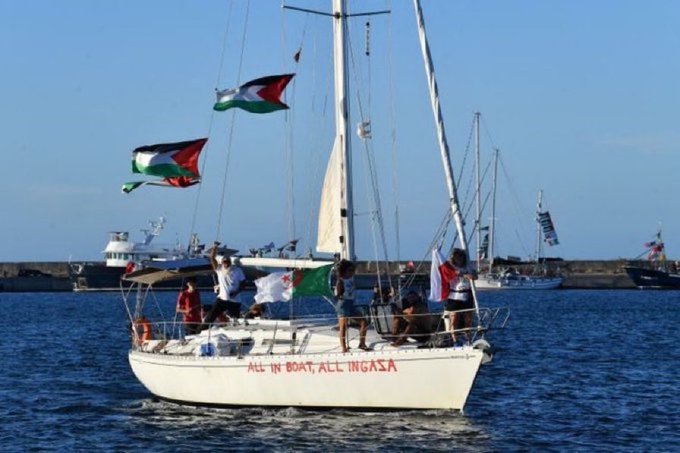 A white sailboat on the water with multiple Palestinian flags and a Spanish flag visible. The boat has red text reading "ALL IN BOAT ALL IN GAZA" on its side. Several people are on deck, and other boats and a harbor are in the background. The Spanish and Israeli flags are shown separately.