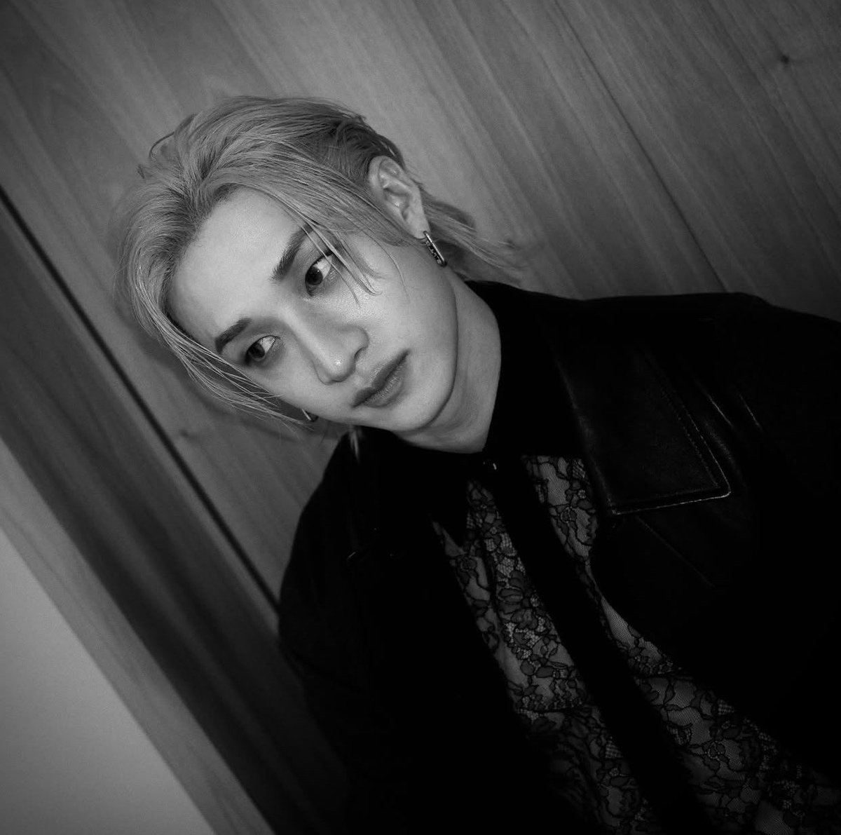 A magnetic essence shines through a poised stance, clad in lace and leather against a wooden canvas, exuding a timeless, shadowy elegance. The monochrome tone deepens the captivating, introspective allure.