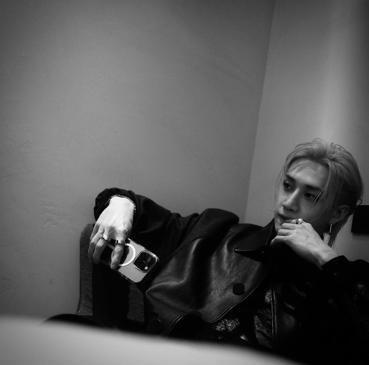 A bold spirit captures a moment of raw intensity, lounging in a sleek leather coat with lace accents, phone in hand against a minimalist wall. The black-and-white filter amplifies the edgy, contemplative allure.