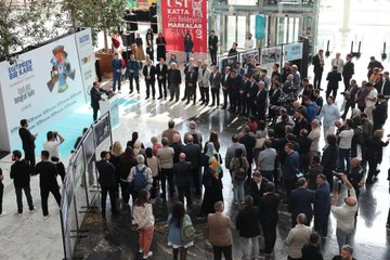 Ali Yalçın speaking at a podium with microphones, addressing a crowd. Yusuf Tekin and İbrahim Yumaklı stand nearby in suits. A large banner displays "Eğitimden Bir Kare Fotoğraf Sergisi" and "Eğitim-Bir-Sen" logo with images. People gather around, some in suits, others casually dressed, near Ankara ATG YHT Gar.