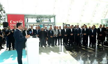 A man in a suit speaking at a podium with a microphone in a spacious indoor area with a modern glass ceiling and greenery. A group of people in formal attire stands behind him, some holding a red ribbon. Banners display "Eğitimden Bir Kare" and logos, with photographs of people in distress visible on display boards. Another image shows a group of people in suits cutting a red ribbon in front of a banner with "Eğitimden Bir Kare" and images of people in distress. Additional photos show men in suits observing photographs on display boards.