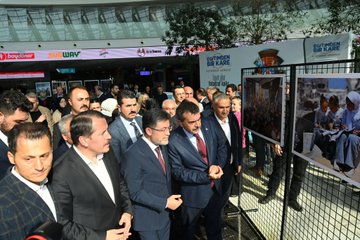 A man in a suit speaking at a podium with a microphone in a spacious indoor area with a modern glass ceiling and greenery. A group of people in formal attire stands behind him, some holding a red ribbon. Banners display "Eğitimden Bir Kare" and logos, with photographs of people in distress visible on display boards. Another image shows a group of people in suits cutting a red ribbon in front of a banner with "Eğitimden Bir Kare" and images of people in distress. Additional photos show men in suits observing photographs on display boards.