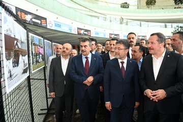 A man in a suit speaking at a podium with a microphone in a spacious indoor area with a modern glass ceiling and greenery. A group of people in formal attire stands behind him, some holding a red ribbon. Banners display "Eğitimden Bir Kare" and logos, with photographs of people in distress visible on display boards. Another image shows a group of people in suits cutting a red ribbon in front of a banner with "Eğitimden Bir Kare" and images of people in distress. Additional photos show men in suits observing photographs on display boards.