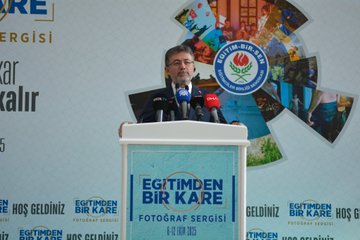 A man in a suit stands at a podium with multiple microphones, including red and black ones, speaking at an event. Behind him, a large banner displays "Eğitimden Bir Kare Fotoğraf Sergisi" with text reading "Hayat akar fotoğraf kalır" and dates "6-12 Ekim 2025." The banner includes logos and images of people and nature. Another image shows a group of people, including men in suits and a woman in a beige coat, cutting a red ribbon in a spacious indoor area with trees and a glass ceiling.