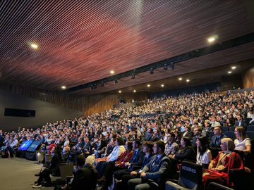 A stage with several people in formal attire, including Yusuful KIRAC, Chairman of the Board and President of Turkish Space Agency, standing at a podium. A large screen displays the Sydney Opera House, text reading "IAC 2026 Antalya," and Yusuful KIRAC\'s name and title. A flag of Türkiye and another flag are visible on stage. The audience is seated in a large auditorium with wooden paneling.