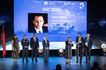 A stage with several people in formal attire, including Yusuful KIRAC, Chairman of the Board and President of Turkish Space Agency, standing at a podium. A large screen displays the Sydney Opera House, text reading "IAC 2026 Antalya," and Yusuful KIRAC\'s name and title. A flag of Türkiye and another flag are visible on stage. The audience is seated in a large auditorium with wooden paneling.