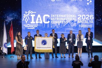 A stage with several people in formal attire, including Yusuful KIRAC, Chairman of the Board and President of Turkish Space Agency, standing at a podium. A large screen displays the Sydney Opera House, text reading "IAC 2026 Antalya," and Yusuful KIRAC\'s name and title. A flag of Türkiye and another flag are visible on stage. The audience is seated in a large auditorium with wooden paneling.