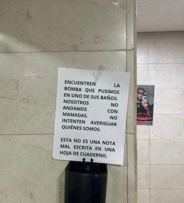 Un trozo de papel pegado a una pared de azulejos con texto negro en español. El texto dice: "ENCUENTREN LA BOMBA QUE PUSIMOS EN UNO DE SUS BAÑOS. NOSOTROS ANDAMOS ANDAMOS NO CON INTENCIONES. AVERIGUEN QUIÉNES SOMOS. ESTA NO ES UNA MAL ESCRITA EN UN HOJA DE CUADERNO." Se ve un cubo de basura debajo del papel. No hay marcas de agua presentes.