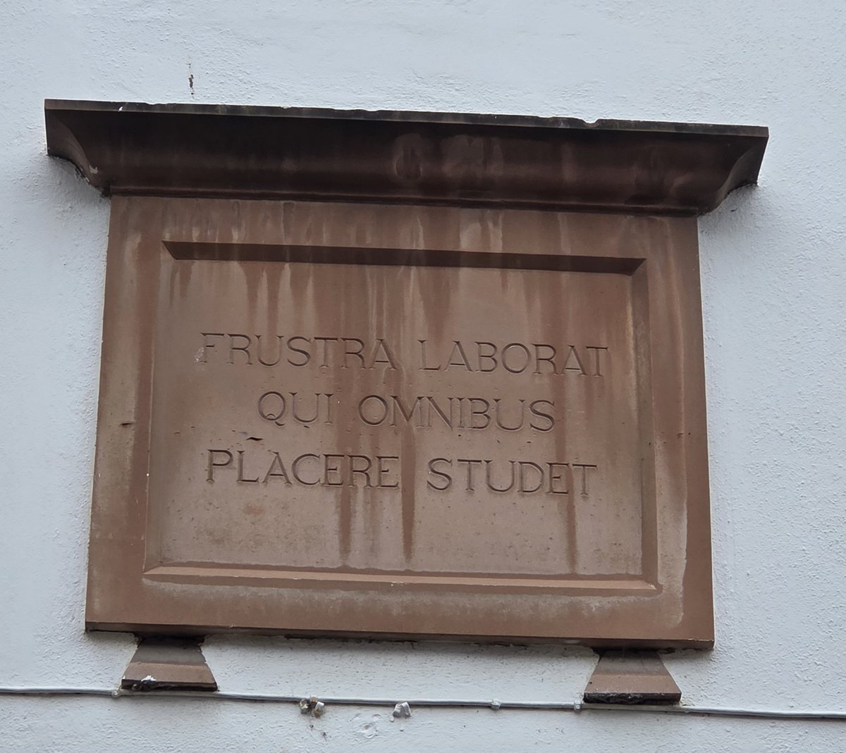 A tongue-in-cheek Latin plaque placed high above a modern urban redevelopment in Penrith, presumably by the architectural firm: "He who tries to please everyone toils in vain".
