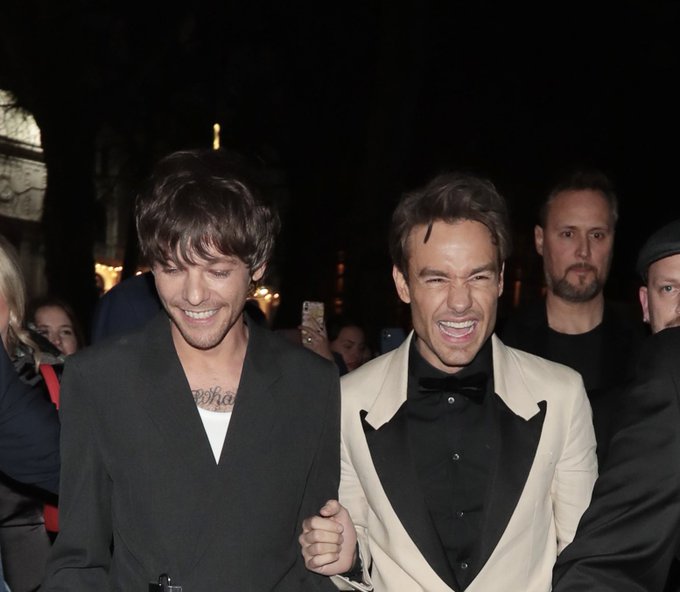 Louis Tomlinson and Liam Payne walking together, smiling. Louis wears a dark suit with a visible tattoo on his chest, and Liam wears a light-colored suit with a black shirt. They are in a crowded outdoor setting at night, with people and lights in the background.