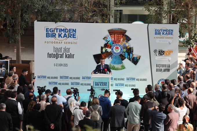 Yusuf Tekin stands at a podium with multiple microphones, delivering a speech. A large banner behind him reads "Eğitimden Bir Kare Fotoğraf Sergisi" and displays photos of educational activities. A crowd of people is gathered, listening attentively. The setting appears to be an outdoor area at Ankara ATG YHT Gar.